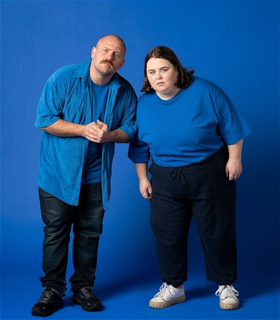 Matt (he/him) is on the left and Taylor (she/her) is standing on the right. They are both wearing blue and are both matching the blue backdrop. They are peering curiously ahead, leaning inward until their shoulders touch.