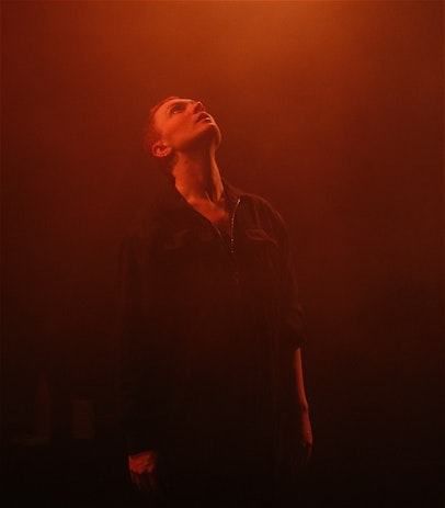 A caucasian person with shaved red hair stands upright, their face is tilted up towards the ceiling. They are wearing a black boiler suit with a silver zipper against a black background. There is smokey haze in the air and they are lit under a warm, orange glow from a stage light.