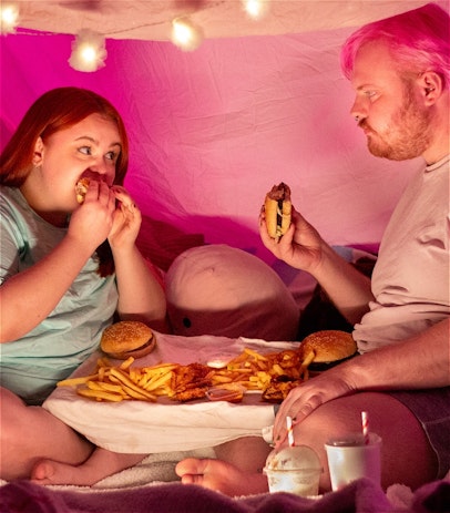 Two fat people sit cross legged facing each other in a pink pillow fort. Between them is a tray filled with french fries and burgers, and drink cups are scattered around them. They are staring at each other while holding burgers and about to take a bite.