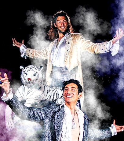 Tom and Josh Burton stand arms outstretched with goofy smiles. They wear sparkly clothes with rings and necklaces and have fake tan on. A toy tiger sits in the middle of them.