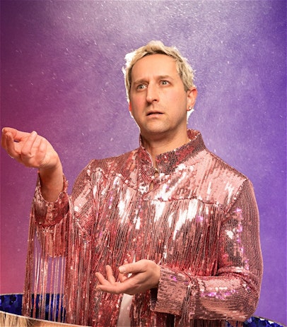 A blonde man wearing a pink sequinned top stares confusedly into the air while bathed in the glow of a spotlight.