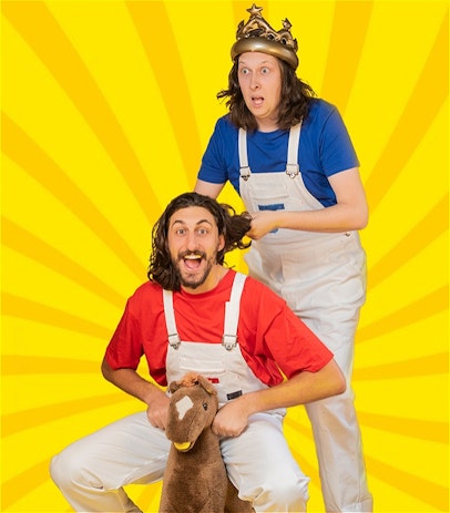 Jero is on a toy rocking horse. Jon is standing behind him looking outwards, wearing an inflatable gold crown. Both are wearing white overalls with a red Tshirt for Jero and a Blue Tshirt for Jon