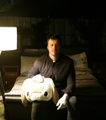 Man sitting on bed outdoors dressed all in black with pastor collar. Plush rabbit head on his knee and floor lamp as light source.