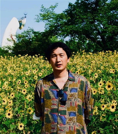 A Japanese man wearing a multi-coloured patterned shirt stands in front of a field of sunflowers.