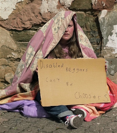 Performer Kathryn Hall is a white woman in her 30s with long brown hair. She is sitting on the ground in front of a rustic wall covered in blankets. She is holding a cardboard sign saying 'Disabled Beggars Can't Be Choosers'.