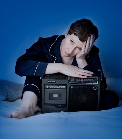 Telia Nevile, a person with short, red/brown hair, sits on a bed with one hand on the boom box sitting in front of her, and her face cradled in the other hand. They look directly into the camera. It appears to be night-time.