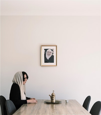 Charaf is sitting at a wooden dining table. She is wearing a black long sleeve dress with a white headscarf as is typical in her tradition while mourning. Her hands are on the table around a small coffee cup. In the middle of the table, a golden brass tray with a traditional coffee pot. The chair on the side of the table facing her is tucked in, a coffee cup identical to the one in her hands is placed on the table in front of the empty, tucked in seat. In the background, a photograph of her deceased grandmother in a golden frame hanging on a white wall.