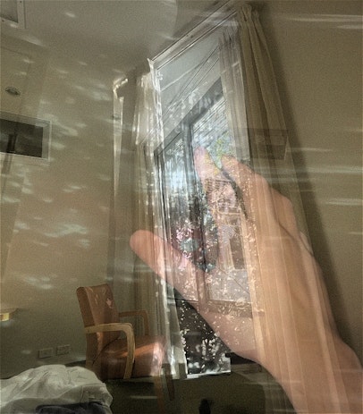 An image of the artist's hand is layered over images of the interior of his hospital room, reaching towards the window. The room is filled with lights that look as though they have all spun together, and the image has a grainy film-like quality to it.