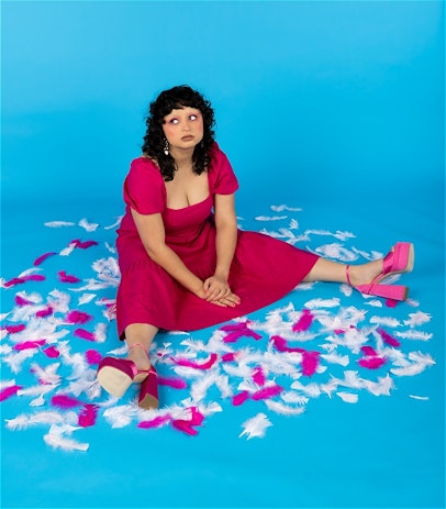 A sky-blue backdrop. Gemma wears a hot pink dress and matching shoes, she is sitting on the floor with her legs out and hands in her lap, surrounded by pink feathers. She is looking a bit defeated and looking off to the side.