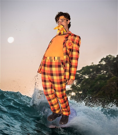 A brown haired man wearing a orange plaid suit is superimposed on top of a surf board riding a long wave at sunset. The full moon is in the sky to the left, and trees line the horizon on the right. The man is leaning backward, and has a half peeled mango sticking out of his mouth.