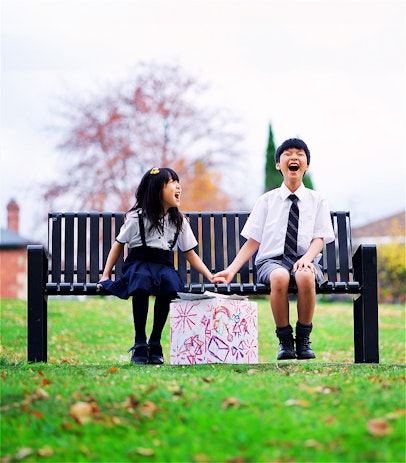 Two children, a boy and a girl, are sitting on a bench in a park. They're holding hands, laughing joyfully together. Positioned in front of them, there's a box brimming with letters