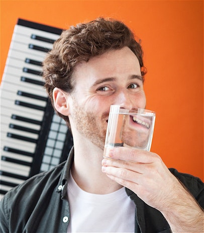 A man in a white t-shirt and green shirt against an orange background. He is smiling and holding a glass of water in front of his mouth, which is mirroring it to face back towards him. A musical keyboard is out of focus behind him.