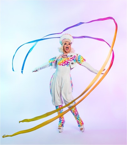 Drag queen Valerie Hex wears a white and rainbow outfit while standing in the middle of swirling rainbow ribbons