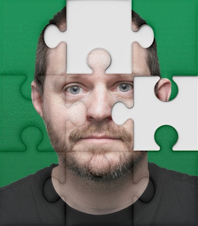 A headshot-photo cut up like a jigsaw puzzle with two pieces missing, in the photo a man looks worryingly at the camera, he is white, mid to late 30s with brown hair and a beard wearing a black t-shirt.