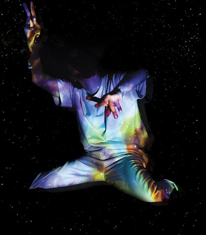 A dark background with small spotty stars. Solo performer Daniel is in a white shirt and pant, lit up in cool blue and yellow galactic lighting. Thier body is contorted, one hand reaching upwards, one towards the camera, knees bent, head sunken into the darkness.