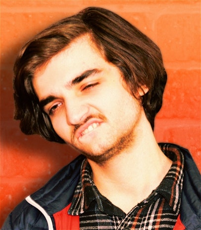 A man with brown short hair and facial hair squints and bites his lip in front of an orange brick wall.