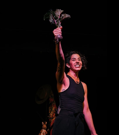 A woman gleefully holds up a beetroot with its stems and leaves attached. She is wearing all black with a traditional Ukrainian coral necklace. A stuffed kangaroo and a Ukrainian scarf are visible in the background.