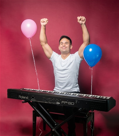 Josh Cake sits behind a piano with a goofy smile and arms raised in triumph. A pink and a blue balloon are floating from the piano either side of Josh. Josh is wearing a plain white T-shirt.