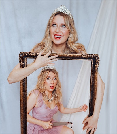 A caucasian, blonde woman in her early 30's is smiling into the distance with a light grey backdrop. She has on glam makeup and wears a tiara . She is clutching a large mirror. Within the mirror is another photo of herself wearing the tiara, a jewelled necklace and a lilac dress. This version of her looks shocked and is holding a knife with blood dripping on the end.