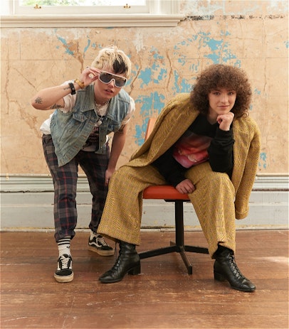 Hannah is looking into the camera with a half-smile, seated on a swivel chair on Rudy's left. Rudy is standing with right hand on knee and left holding white-framed sunnies as they lean into the camera. Rudy wears a denim vest, check pants and sneakers, while Hannah has a big-haired 80s vibe in an oversized tweed suit. It's giving 'Breakfast Club'