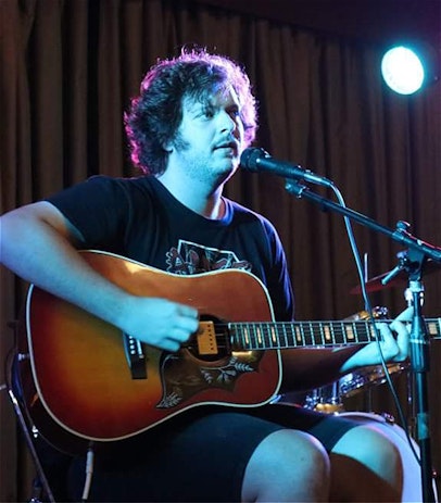 A performance picture of a young man sitting in a chair playing an acoustic guitar with a microphone set up in front of him.