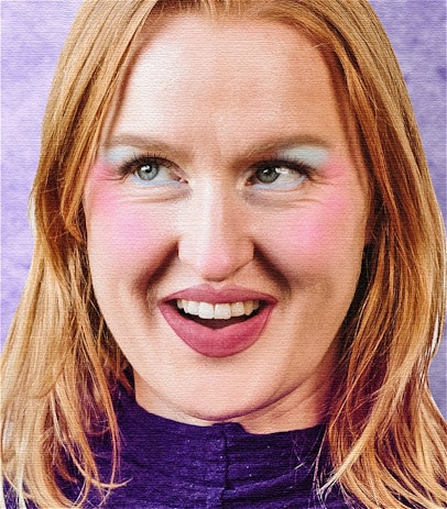 A close up photo of a woman smiling and looking up to her left with her eyes with red hair, she is wearing a purple top in front of a purple backdrop, she is wearing blue eyeshadow, pink blush and lip liner.