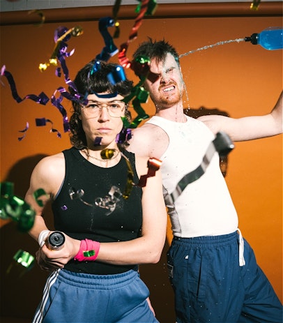 A young person shoots confetti towards the camera. Their face is seen through the confetti, looking at the camera. They are wearing glasses and short brown hair with a mullet. Another young person behind is squirting a bottle of blue liquid into their face. The liquid is splashing into their face and they have their eyes shut. The have short brown hair.