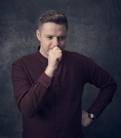 Image of Tom Ballard holding his hand in a first in front of his mouth, with his other hand on his hip. He is wearing a maroon button up shirt against a grey background.