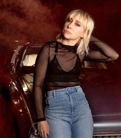 A woman with blonde hair, dressed in a sheer black top and jeans, leans on the front of a car. She is standing with one hand behind her head, the other hanging by her side. It's night time and there is a red mist surrounding her and the car.