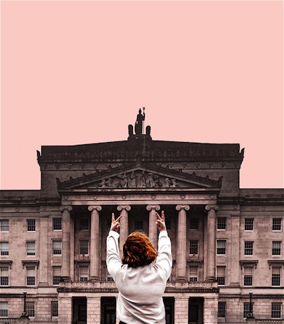 A young woman facing away putting two fingers up to Stormont (Northern Ireland's government building).