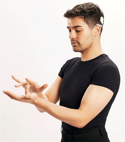 A tall man with olive skin and has dark features, such as dark-trimmed beard and dark wavy hair. He is signing 'middle' in Auslan (Australian Sign Language) which the left hand is flat, and the other hand has the middle finger placing onto the flat hand which is the definition of that is 'middle' in Auslan. There is a white cochlear implant hooked onto his ear. The man is wearing a tight black tee tucked in a high-waist black business pants.