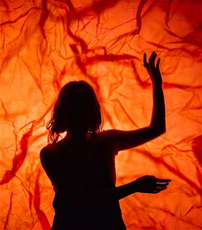 A woman stands in front of a screen that features a firey projection through crumpled cellophane. She holds up her arms like a matador, or a dancer.