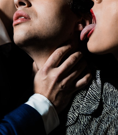 A close up shot of three people. You can only see their shoulders and the bottom halves of their faces and there are shadows across most of the image. On the far right, a person with red lipstick on has their tongue out, about to lick the face of the person in the middle. The person in the middle is wearing a striped navy and white shirt, and they have their face tilted up with a look of both pain and pleasure. On the far left, a person in a blue suit is whispering in the ear of the person in the centre. They also have their hand around the throat of the person in the centre.