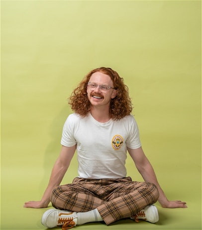 Wide up shot of Nick smiling. He's got long, red, curly hair, a moustache and wearing glasses. In a white shirt and tan, checked pants , he's sitting with both legs crossed and his hands on the ground.