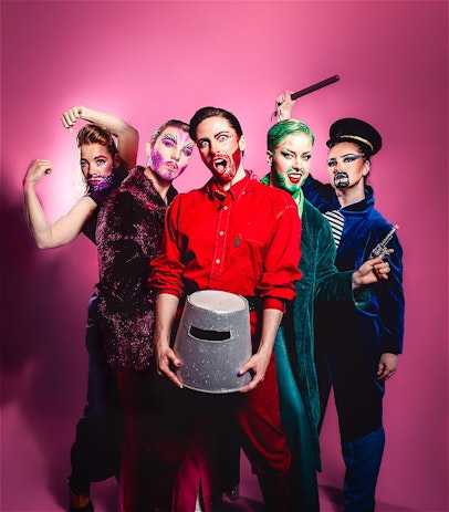 Pink back drop with five drag kings posed in front. Each individual has a monochromatic colour scheme with glittery beards. The person in front is holding a Ned Kelly helmet and has a shocked expression.