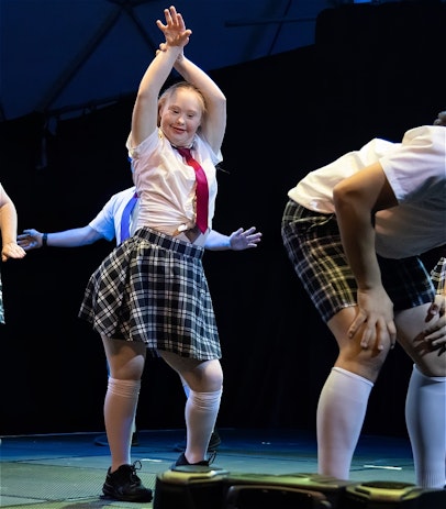 A group of dancers in school uniforms. Long white socks, navy and white, checked netball skirts, white shirts, and red neck ties.
The dancer in focus is holding their hands above their had and smiling.