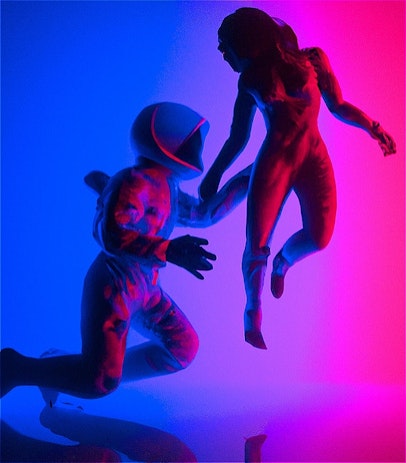 On a background that fades from deep blue to a bold pink colour an astronaut is seen reaching for a floating female figure whose face is obscured by shadows.