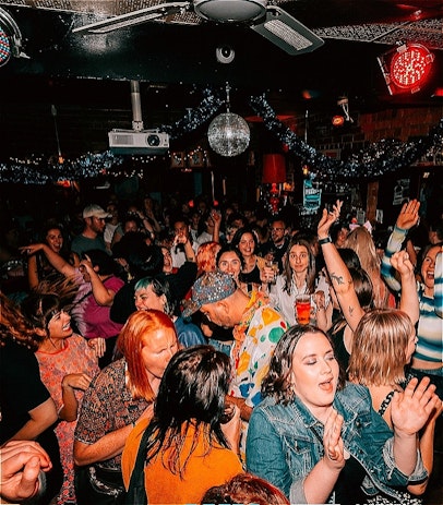 A colourful crowd of people, most of them femme-presenting, on a dance floor under a disco ball.