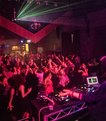 A room full of people dancing, they're bathed in a pink light and a DJ console can be seen at the bottom of the image.