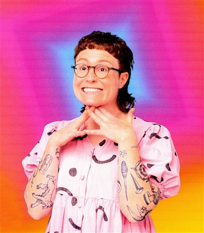 Scout Boxall has a mullet and glasses. They wear a pink shirt against a blue background and stand at a forty-five degree angle looking away from the camera with a smile.