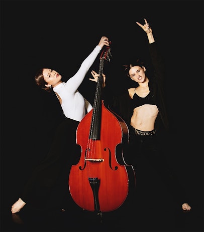 Two dancers are posed either side of a double bass. The dancer on the left is leaning back in a white top with a low bun. The dancer on the right is faced forward in a black bra. They have spiked hair and are making a rock and roll gesture with their hand.