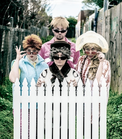 Four people in outlandish outfits peer over a white picket fence