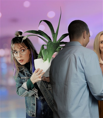 A young woman with purple hair and glasses is hiding very badly behind a plant. She is spying on a beautiful blonde wearing a pink dress, who is smiling at a man in a blue shirt.